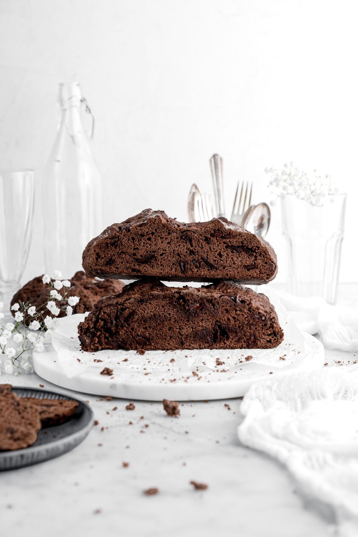two halves of chocolate soda bread stacked on top of each other on marble surface with white flowers and crumbs around and another loaf of chocolate soda bread behind.