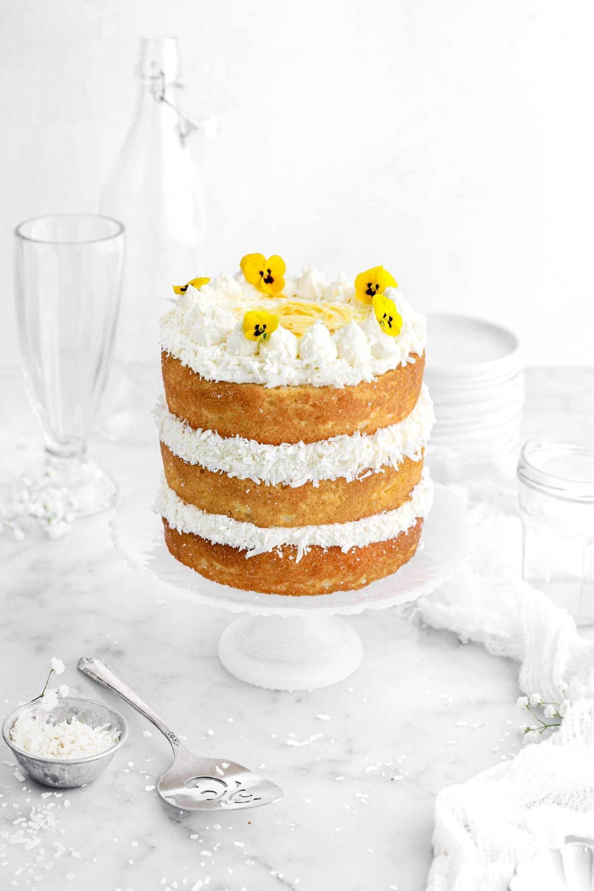 three layers of naked lemon coconut cake with yellow flower son top on a white cake plate, a cake knife, bowl of shredded coconut, and white flowers around on marble surface.