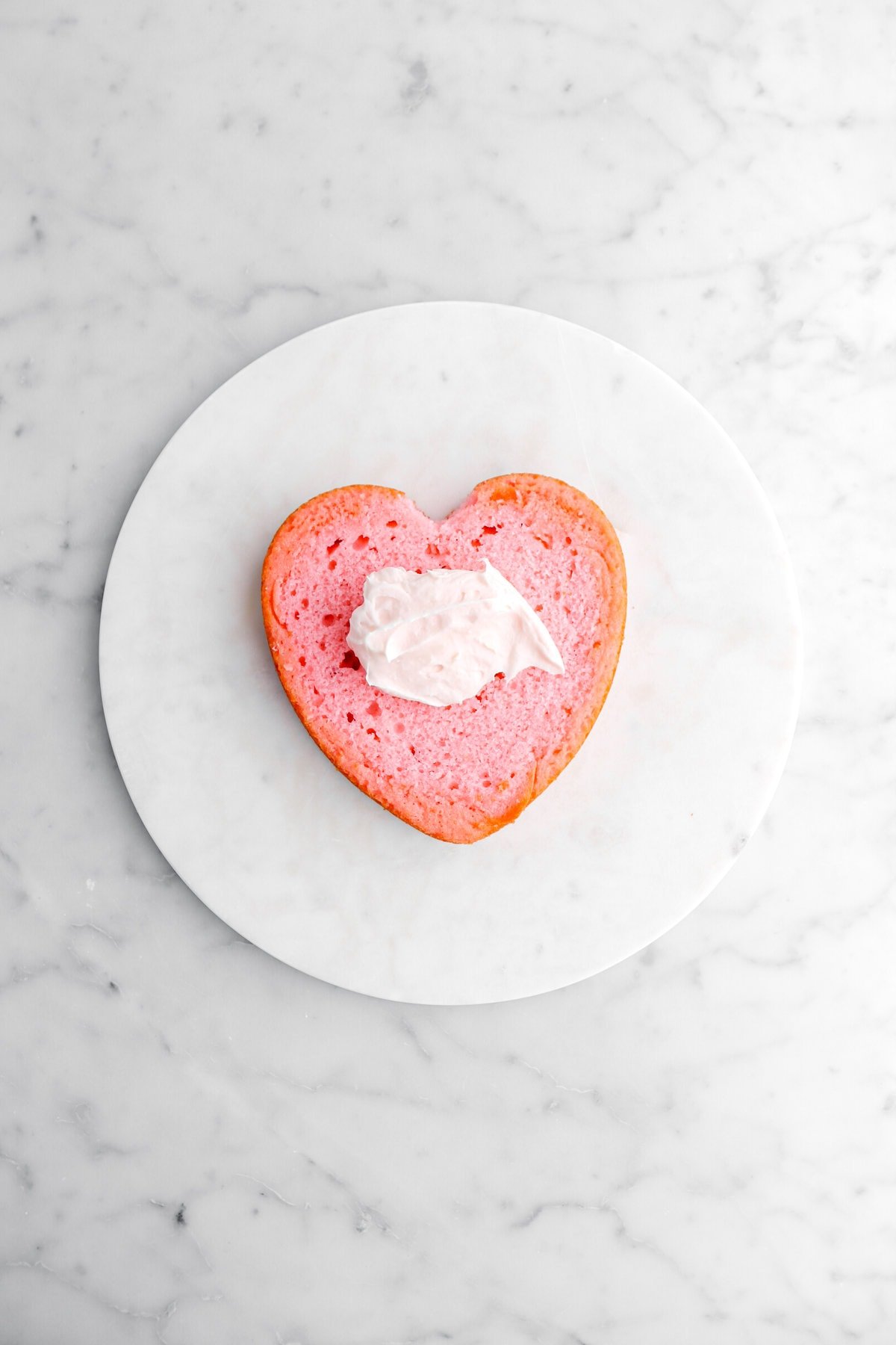 pink heart shaped cake on marble surface with pink cream cheese frosting dolloped on top.