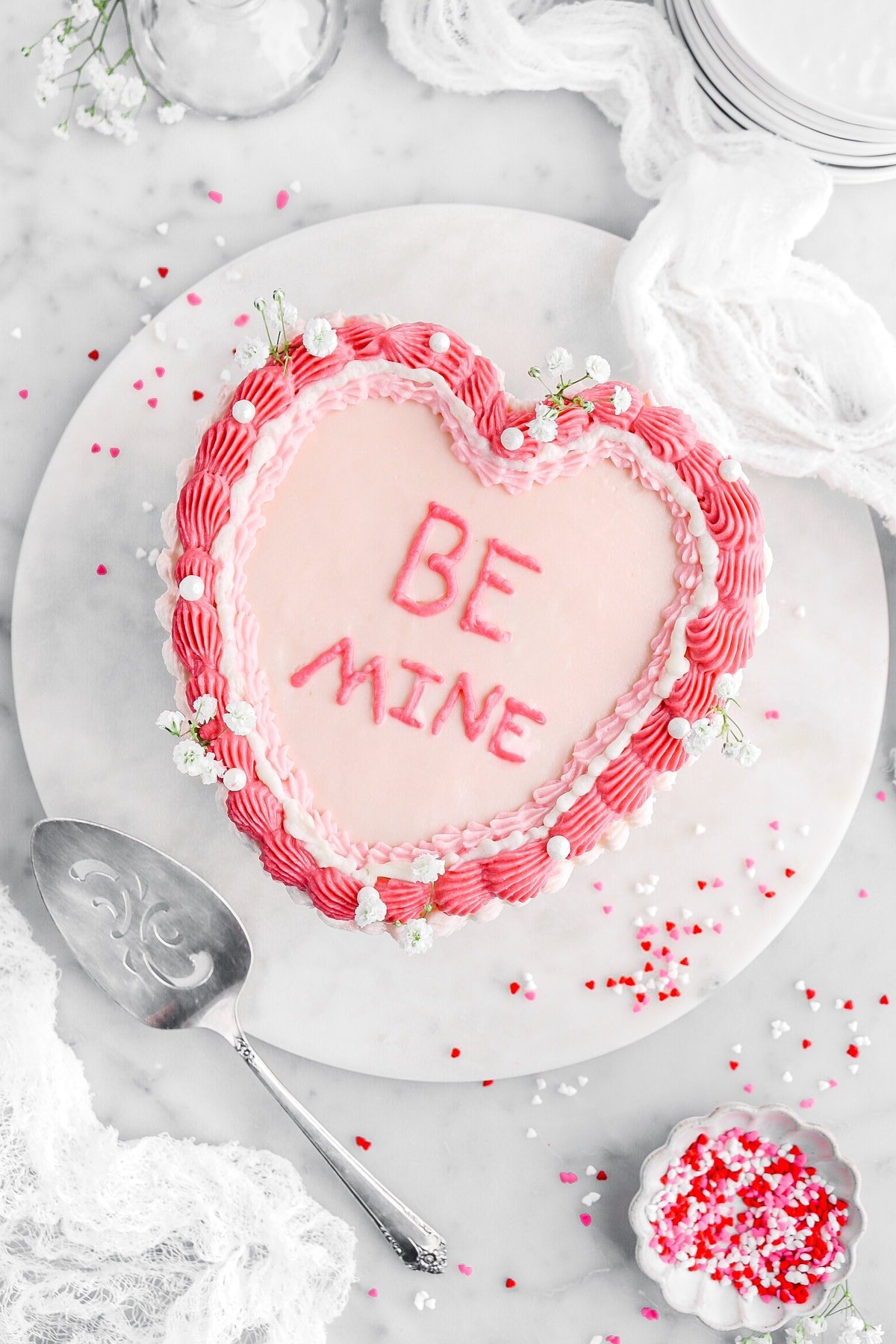 overhead image of decorated pink heart shaped cake with "be mine" written on top with white cheese cloth, heart shaped sprinkles, and a cake knife beside.