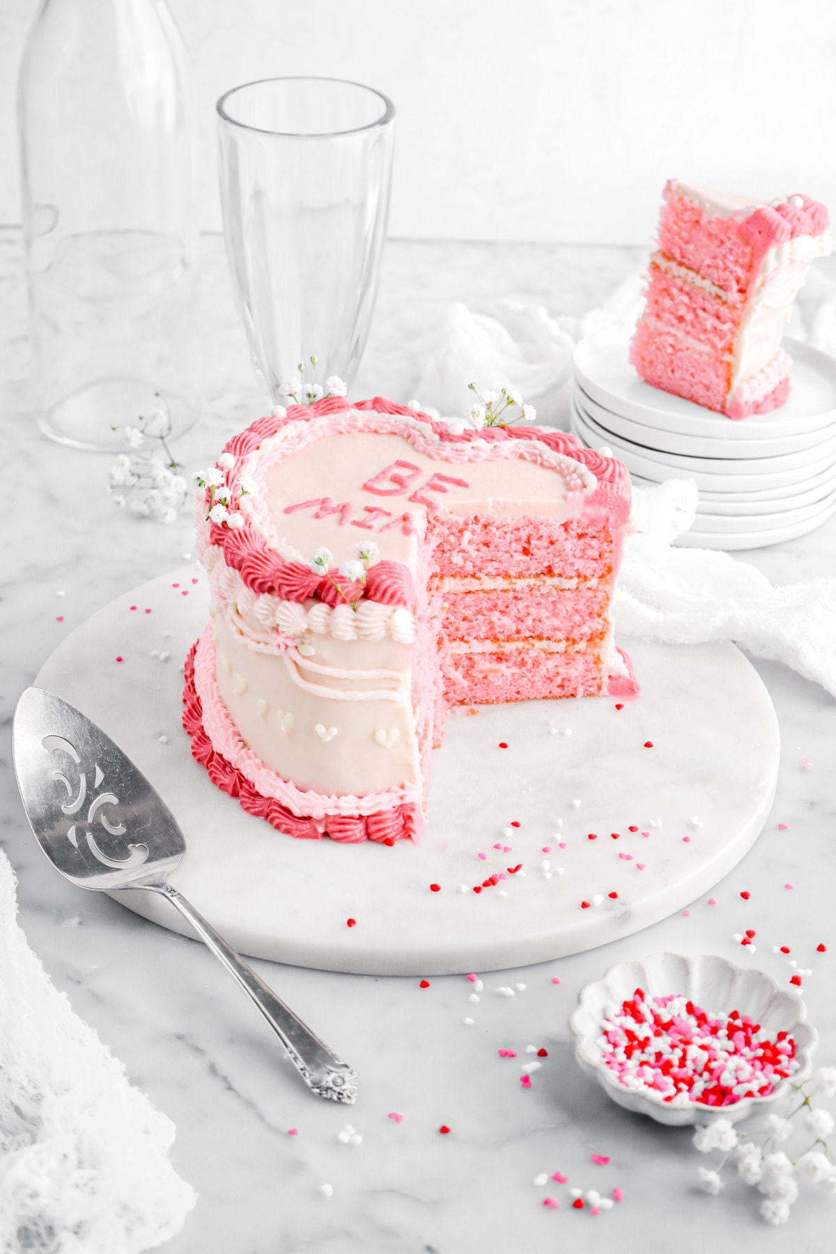 pulled back angled image of cut heart shaped pink velvet cake on marble cake plate with slice of cake behind on stack of white plates.