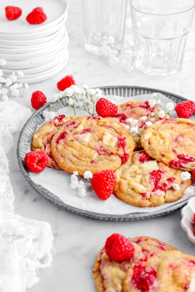 angled close up of raspberry white chocolate cookies on parchment lined plate with fresh raspberries and white flowers around, stack of plates, and two empty glasses behind on marble surface.