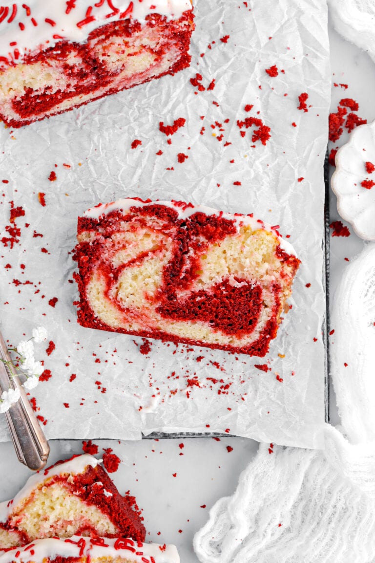 overhead image of slice of red velvet swirl cake on parchment paper with loaf cake above and two more slices of cake below with red cake crumbs scattered around.