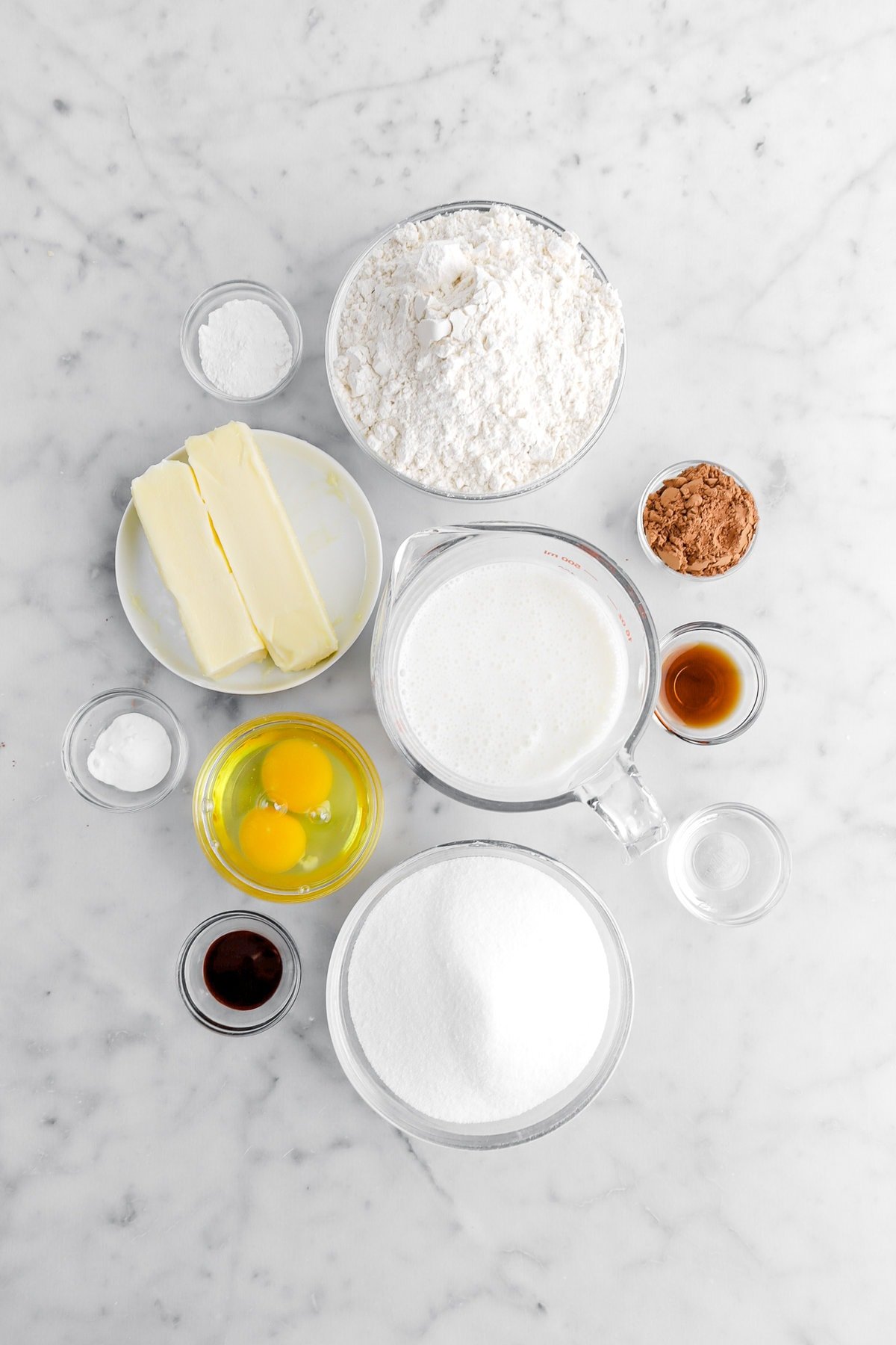 flour, baking powder, butter, buttermilk, cocoa powder, vanilla, vinegar, sugar, red food dye, eggs, and baking soda on marble surface.