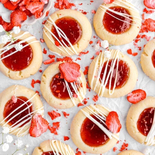 pulled back overhead image of strawberry thumbprint cookies on parchment paper with white flowers and dried strawberries around.