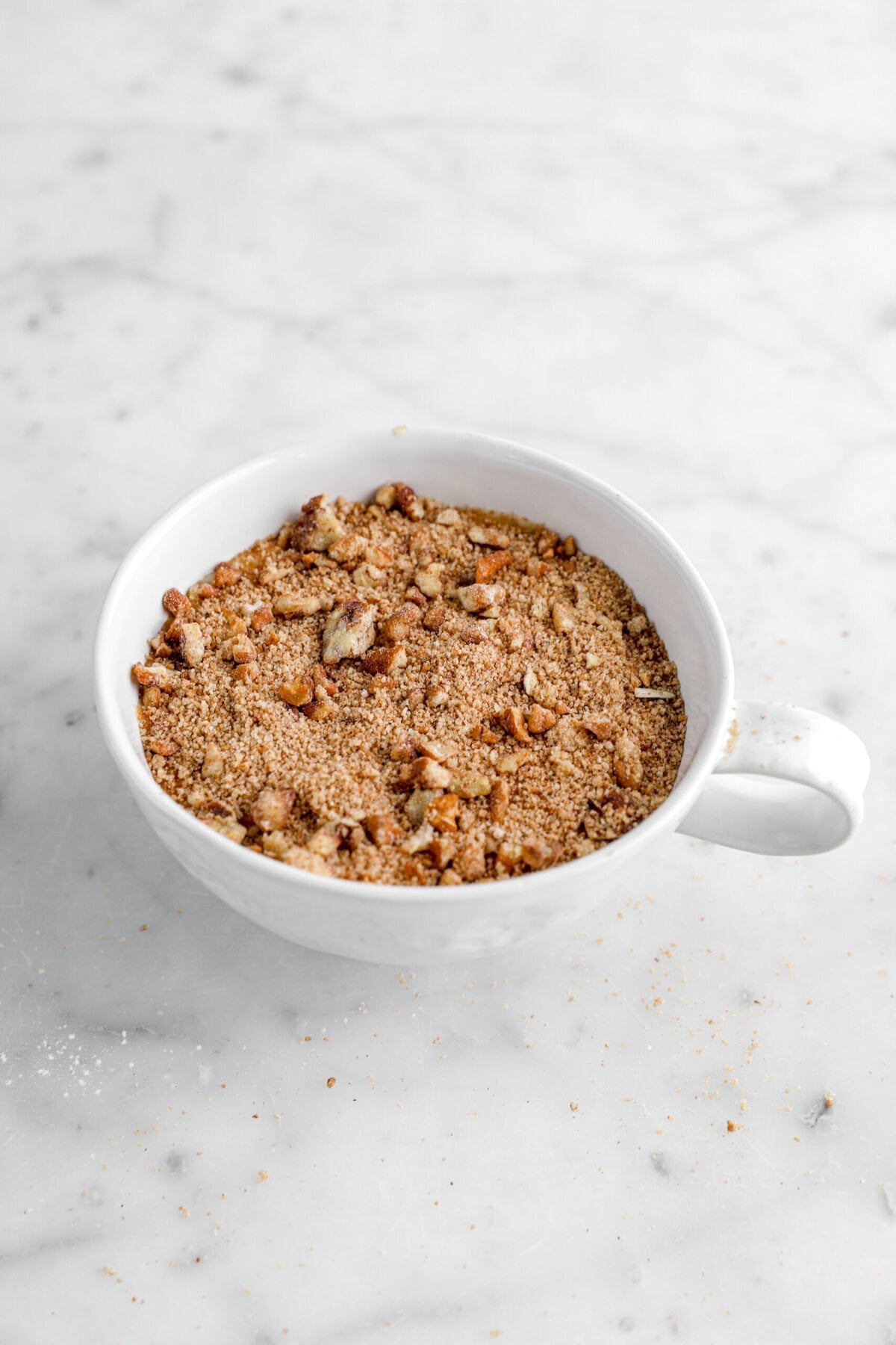 Close-up of crumbly crumble topping in a white cup on marble surface.
