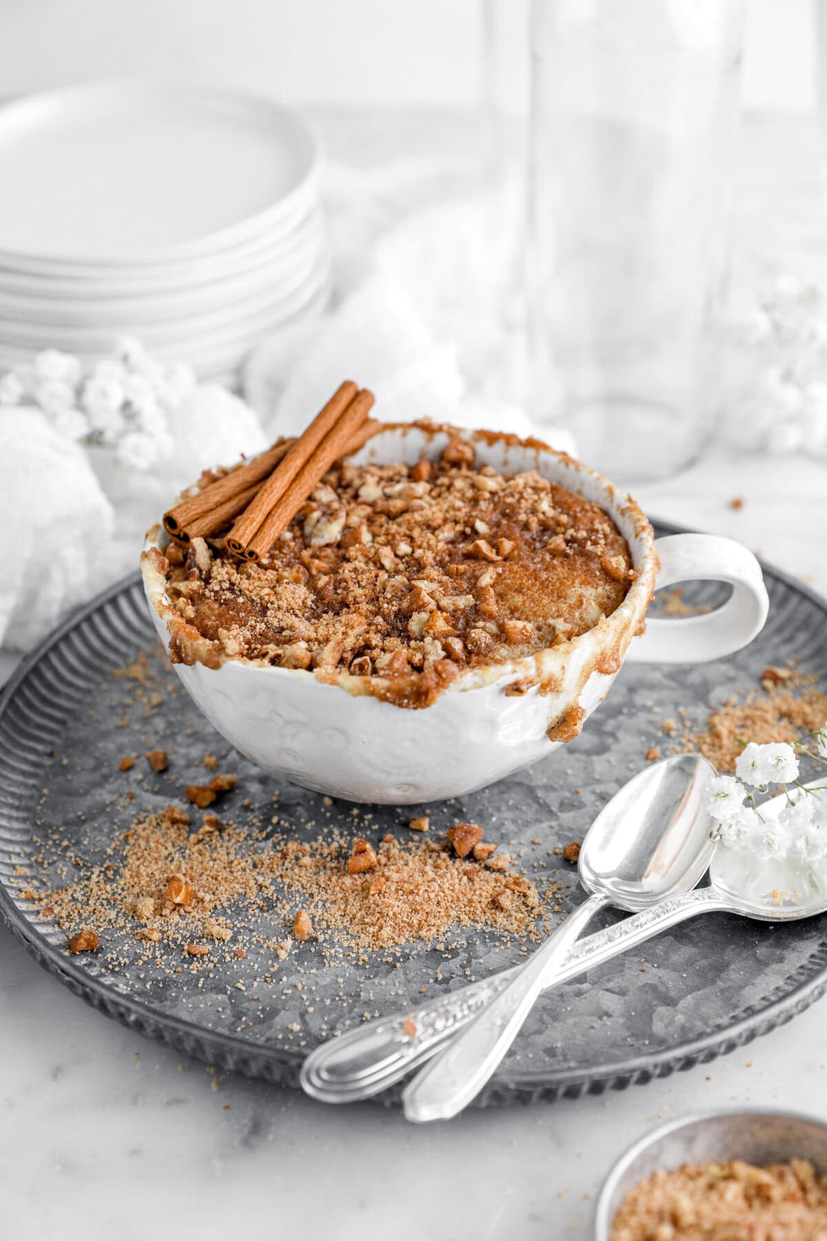 cinnamon coffee cake in white mug with two sticks of cinnamon on top, all on a metal plate with streusel topping sprinkled around and two silver spoons beside mug.