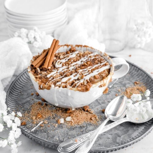 cinnamon coffee cake with icing drizzled on top in a white mug atop a metal plate with two spoons and small white flowers around.