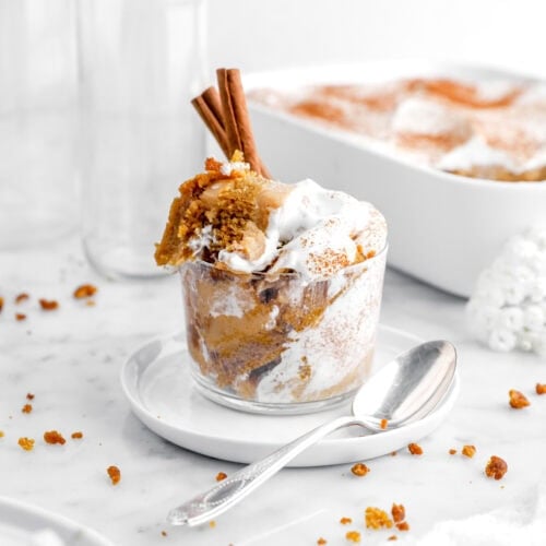 pumpkin bread pudding in glass cup on a small white plate with spoon beside and casserole of pudding behind.