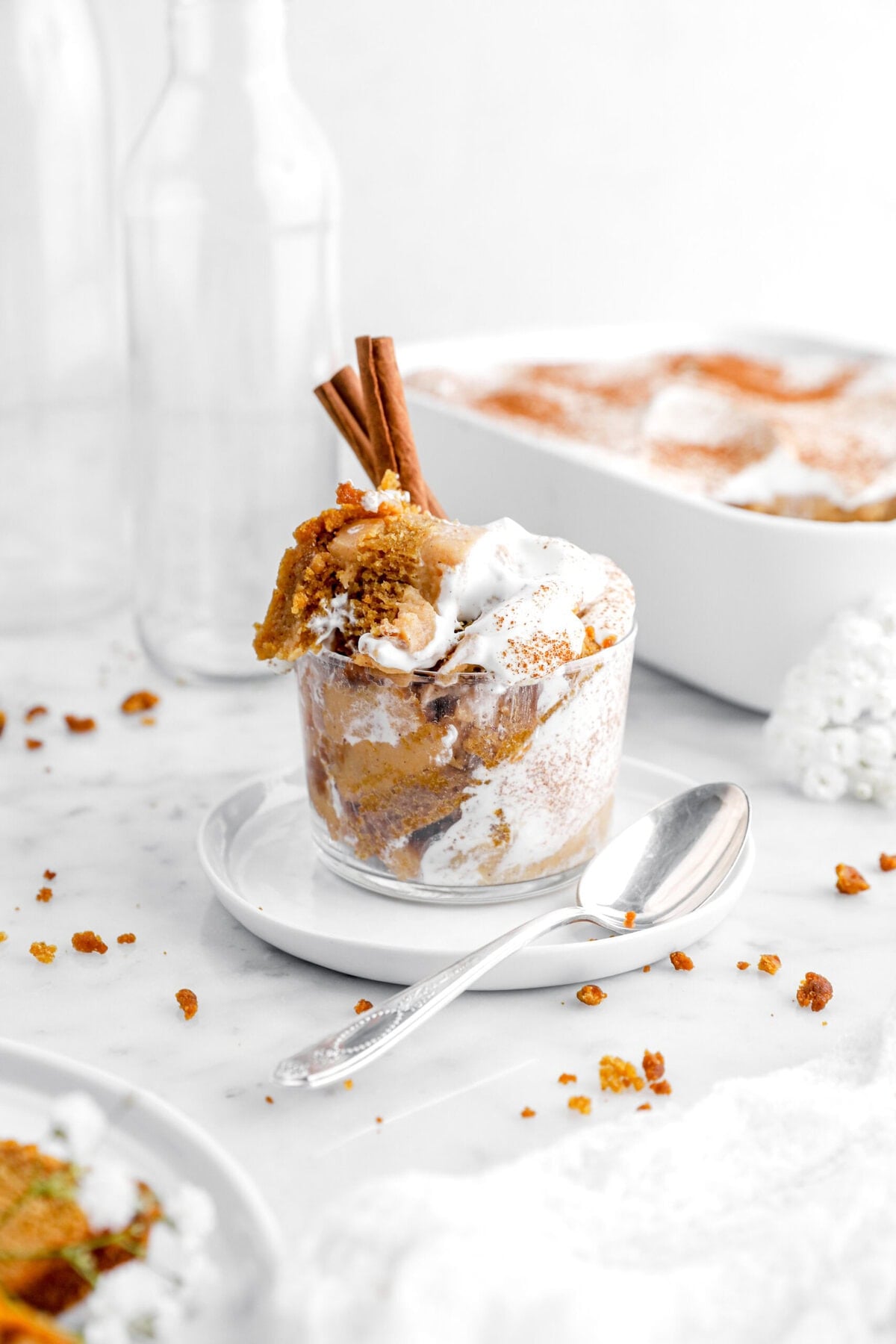 pumpkin bread pudding in glass cup on a small white plate with spoon beside and casserole of pudding behind.