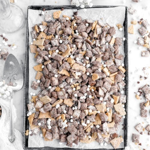 s'mores muddy buddies on lined pan with the snack mix scattered around on marble surface.